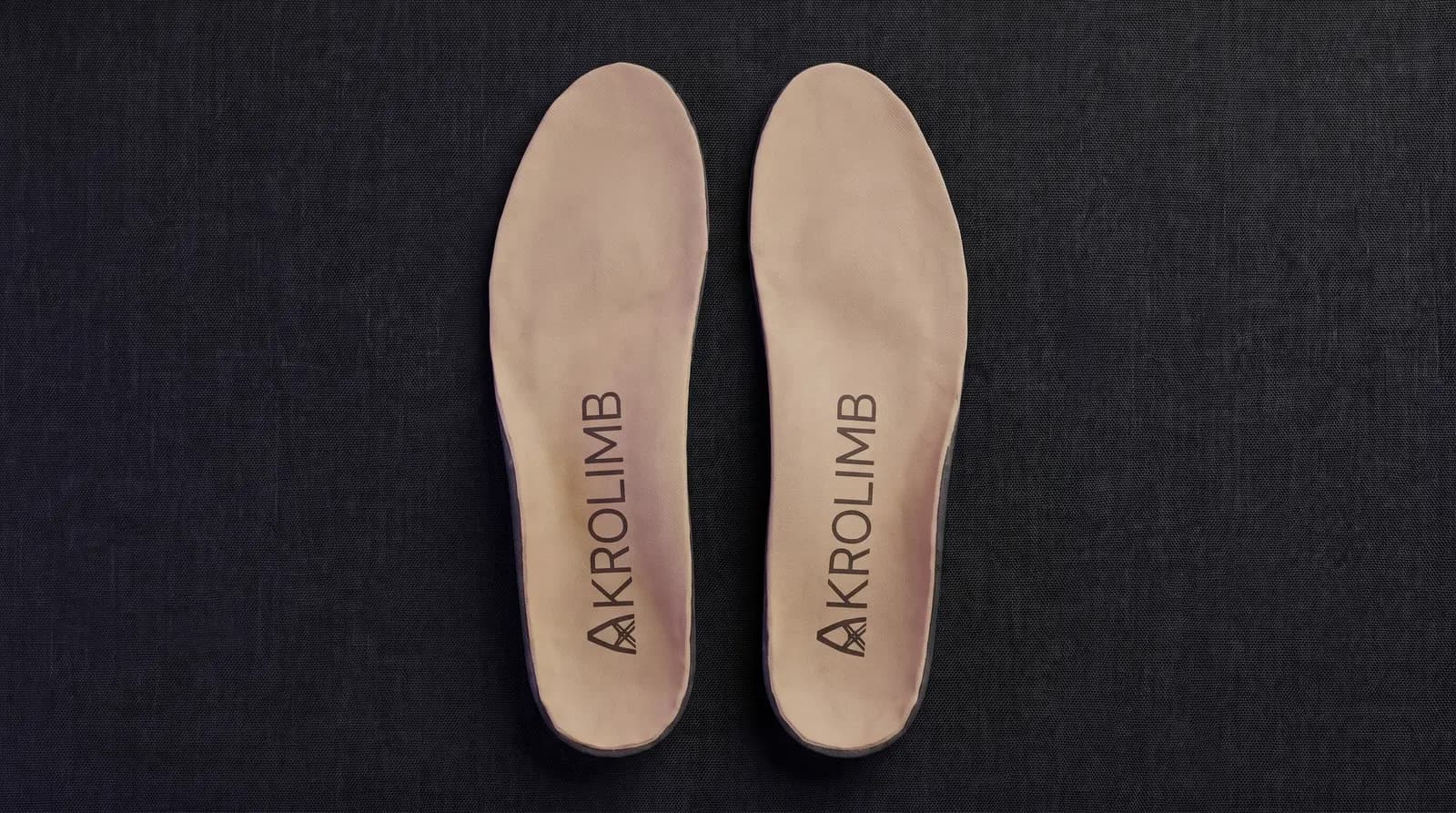 A pair of Akrolimb insoles seen from above, each printed with the AKROLIMB wordmark, on a dark linen surface